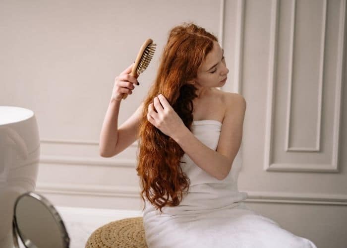 A woman wrapped with a towel brushes her long auburn hair extensions with a wide-tooth hair brush in a living room